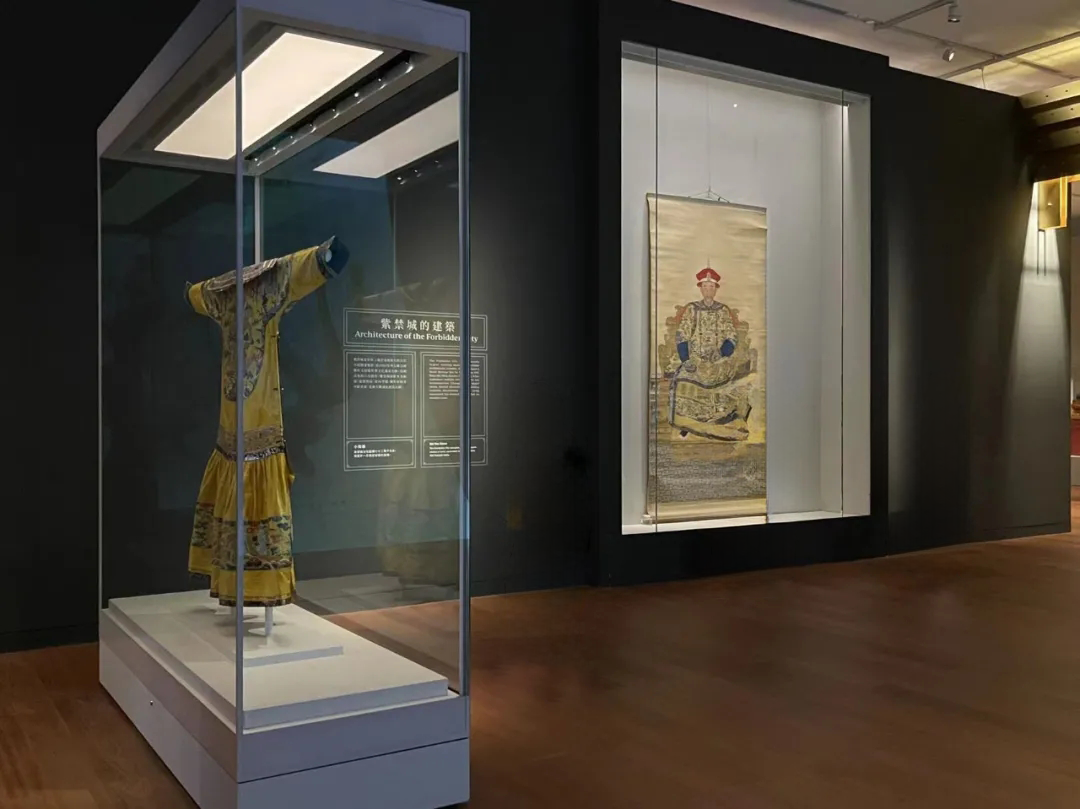 Museum gallery with a glazed garment display case, overhead lighting and an adjacent hanging portrait display