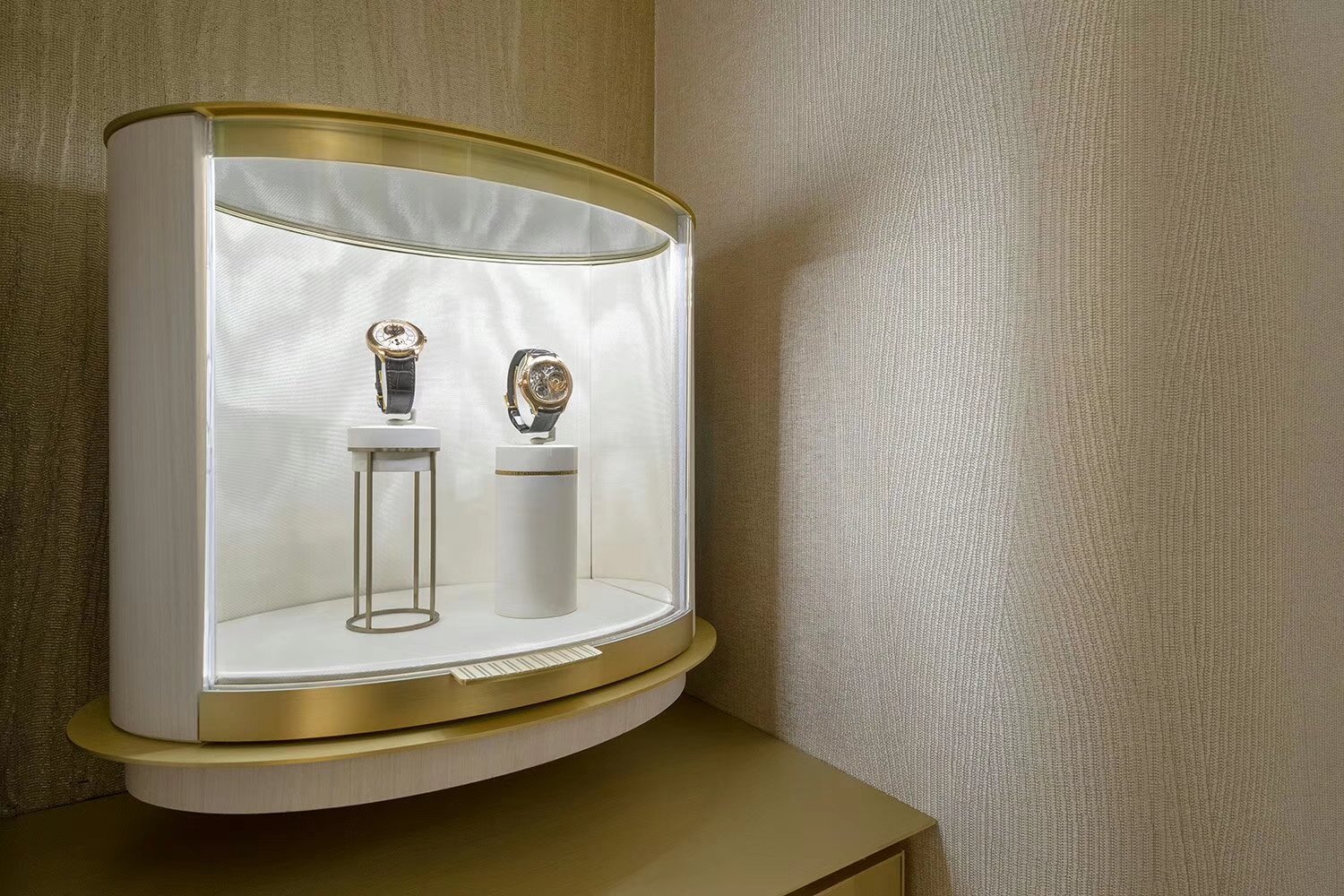 Two wristwatches on raised white display stands inside a curved glass showcase with a gold-toned frame